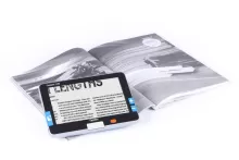 siyah-beyaz bir dergi sayfasının üzerine yerleştirilmiş Optelec Compact-7 HD taşınabilir büyüteç cihazı görülüyor. ekranında "LENGTHS" kelimesinin büyütülmüş hali yer alıyor. Cihazın kenarlarında mavi renkte büyütme seviyesinin ayarlanmasını sağlayan artı ve eksi düğmeleri var, Arka planda açık bir dergi sayfasında modern bir yapı fotoğrafı bulunmakta.